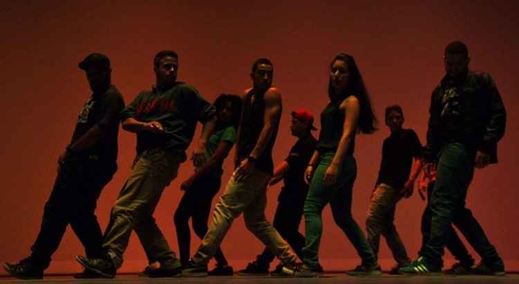 Bailarinos do grupo de dança Tekamül durante apresentação. Nove dançarinos aparecem enfileirados diante de um fundo vermelho.