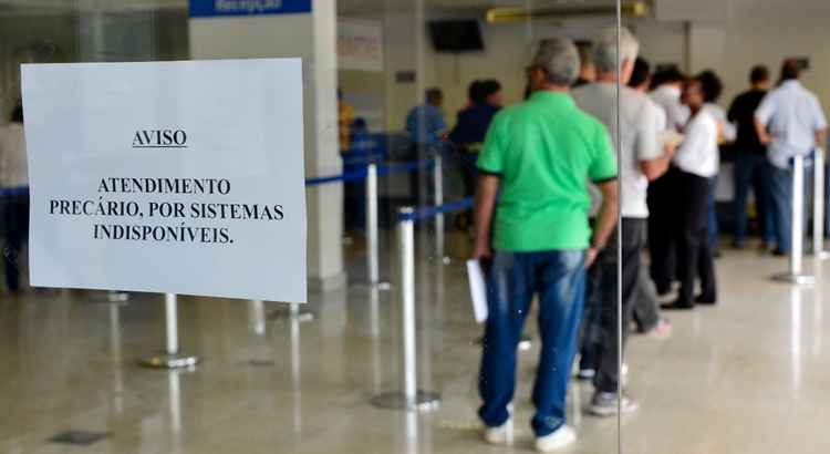 Pessoas em fila no interior de agência do INSS. Em primeiro plano, um aviso onde se lê: Atendimento precário por sistemas indisponíveis