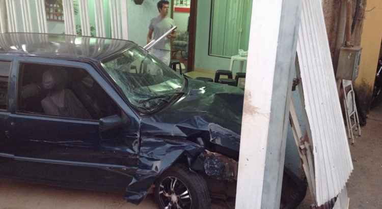 Carro azul amassado no interior de uma casa com paredes verdes e um homem observando a cena