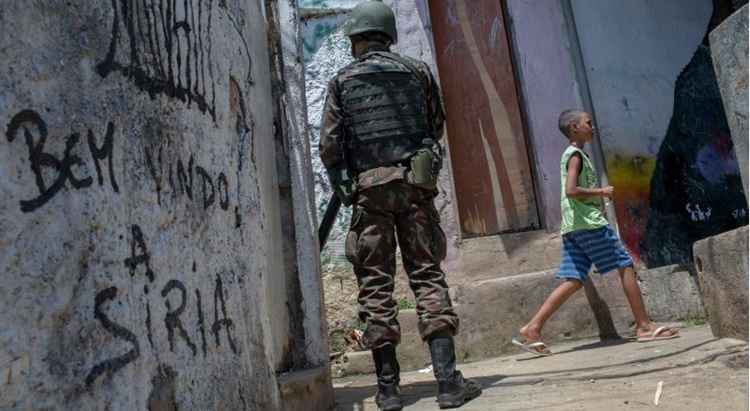 Militar patrulha a Rocinha enquanto criança passa caminhando à sua frente; na parede de uma casa, a inscrição: 'Bem vindo à Síria'