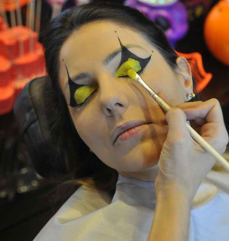 Mulher branca deitada sob uma cadeira de salão para iniciar uma maquiagem para a festa de halloween. A maquiadora estava passando sombra metálica amarela nas pálpebras da moça.