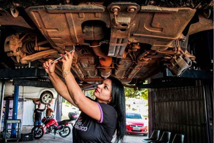 Em baixo de um carro suspenso, mulher em pé, usa camiseta preta, calça jeans e sandália de salto, enquanto trabalha na mecânica do carro