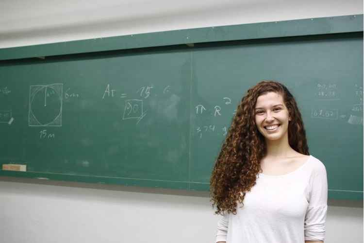 Jovem de cabelos longos, cacheados e blusa branca sorri. Ao fundo, um quadro negro de sala de aula