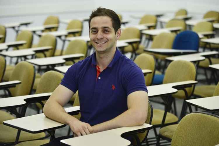 Jovem de camiseta polo escura, sentado em uma carteira de colégio, posa de braços cruzados e sorri. Há outras carteiras em volta dele, sem ninguém