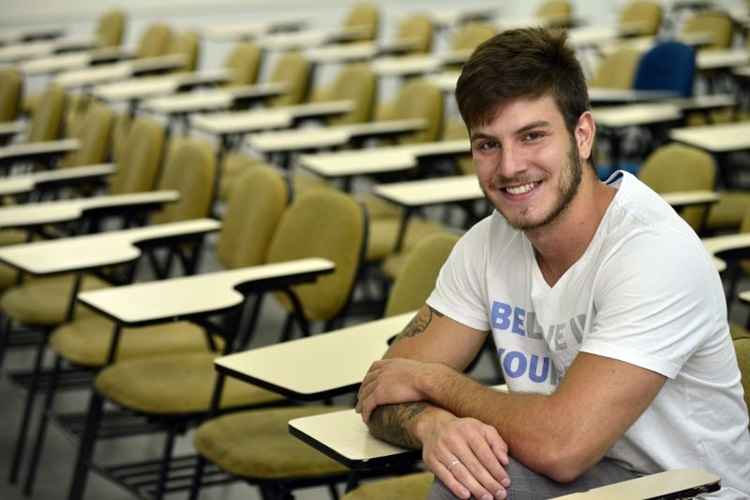 Jovem branco, de camiseta branca, sentado em uma carteira de colégio, posa de braços cruzados e sorri. Há outras carteiras em volta dele, sem ninguém