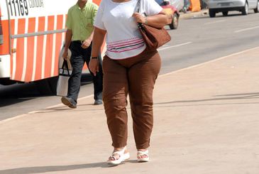 Mulher acima do peso, com camisa branca, calça marrom e sandália branca