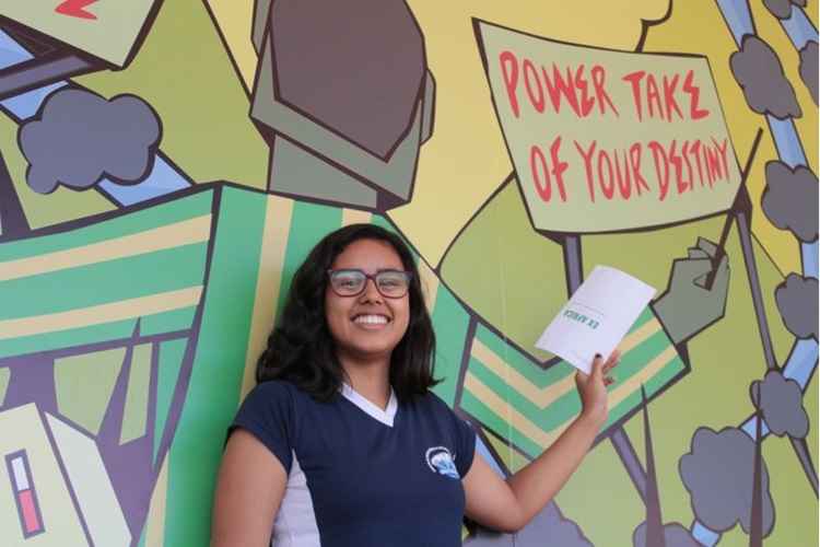 Jovem mulher, de óculos e camiseta escura com símbolo do colégio, sorri em frente ao mural, no qual se lê %u201CPower take of your destiny%u201D