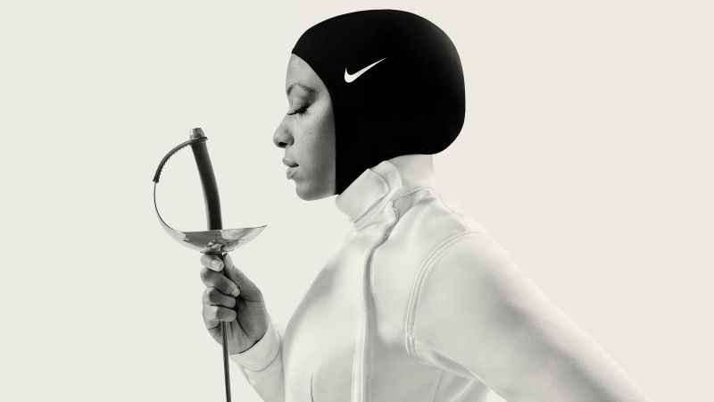 Uma mulher muçulmana de perfil, com um hijab esportivo da Nike preto, uma roupa branca de esgrima, segurando uma espada de esgrima