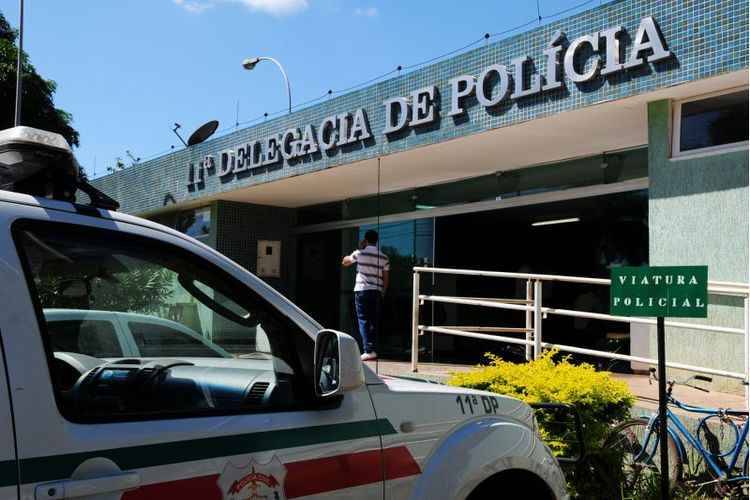 Prédio verde com texto 11ª Delegacia de Polícia escrito no alto; parte de carro branco, com faixas verde e vermelha, sirene no alto e logo da Polícia Civil