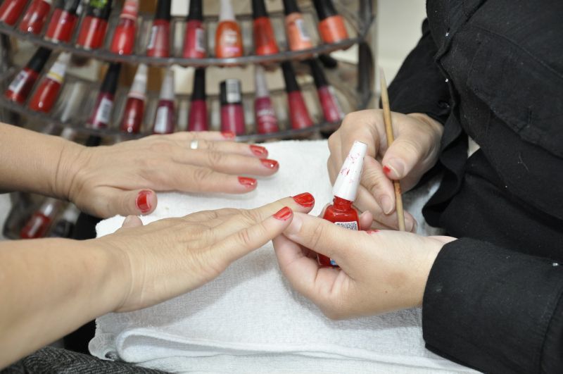 Mãos com unhas com esmalte vermelho; esmaltes; itens de manicure; toalha branca; mão com aliança; casaco preto