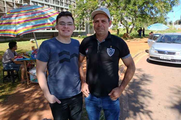 Pai e filho aguardando o início das provas. O jovem está vestido de camisa azul e calça preta e, o pai, de camisa preta e calça azul