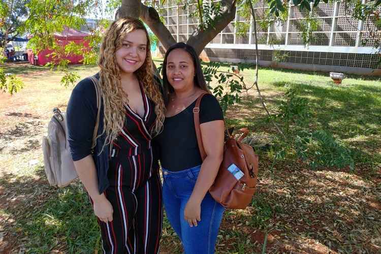 Duas jovens embaixo de uma árvore. Uma vestida de macacão com listras e, a outra, usando blusa preta e calça azul