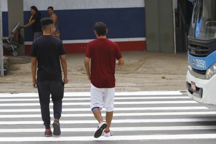 Dois homens atravessando faixa de pedestres; ônibus azul e branco; cone de trânsito; mulher com carrinho de bebê ao fundo; parede azul, branca e vermelha