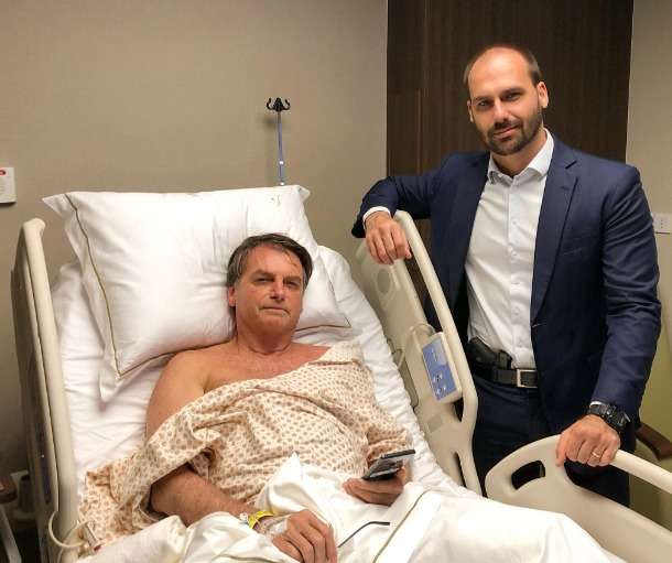 Bolsonaro em uma maca e Eduardo Bolsonaro do lado com uma arma na cintura