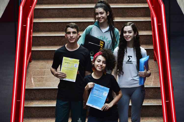 Três meninas e um menino posam em uma escada com corrimão vermelho. Eles estão com a feição séria e seguram cadernos de provas passadas do Enem. Eles são alunos do ensino médio do Centro Educacional Sigma.