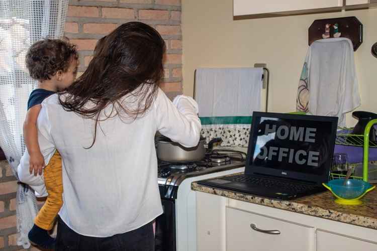 Mulher na cozinha de costas usando uma calÃ§a preta de blusa branca com uma crianÃ§a no colo