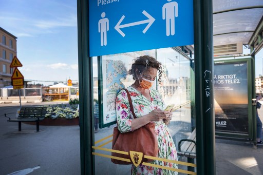 Uma mulher usa máscara facial enquanto espera no ponto de ônibus com um sinal de informações pedindo às pessoas para manter distância social devido à pandemia de coronavírus COVID-19 em 26 de junho de 2020 em Estocolmo, Suécia.